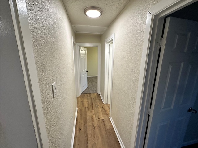 hall featuring light wood finished floors, attic access, a textured ceiling, and a textured wall