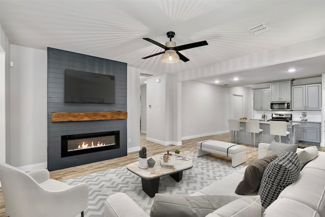 living area featuring a large fireplace, a ceiling fan, visible vents, baseboards, and light wood finished floors