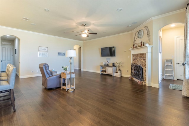 living area featuring arched walkways, crown molding, dark wood-type flooring, a brick fireplace, and ceiling fan