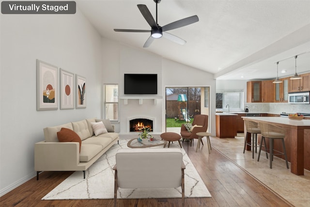 living room featuring baseboards, a ceiling fan, light wood-style flooring, a lit fireplace, and high vaulted ceiling