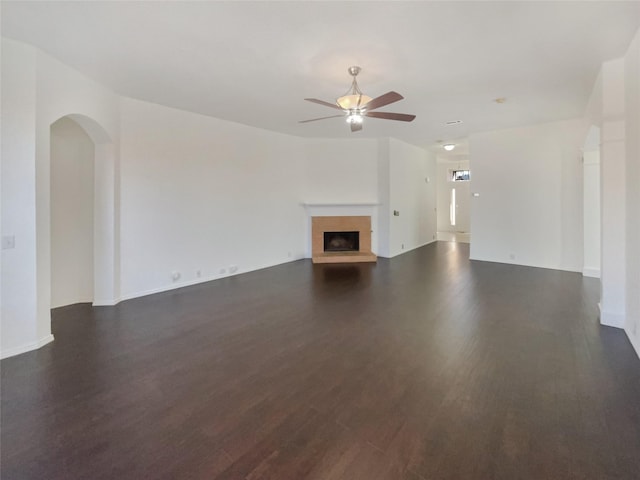 unfurnished living room featuring a fireplace with raised hearth, ceiling fan, dark wood-type flooring, and arched walkways