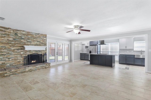 unfurnished living room featuring a stone fireplace, a sink, visible vents, a ceiling fan, and crown molding