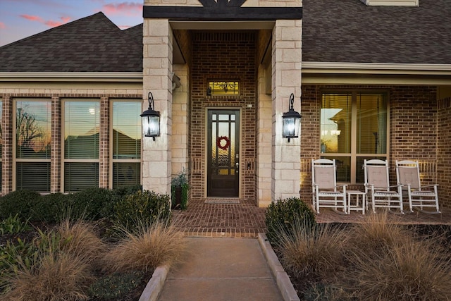 view of exterior entry featuring stone siding, covered porch, brick siding, and roof with shingles