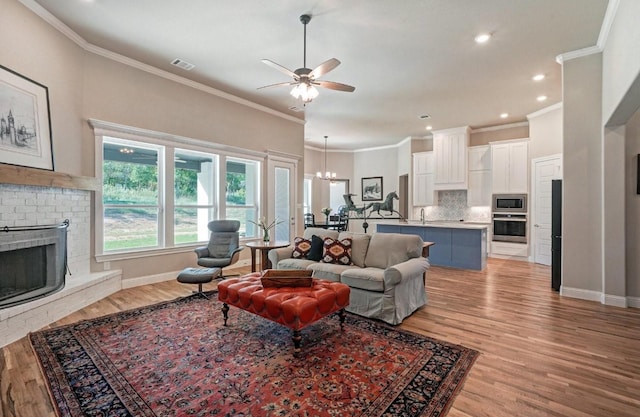 living area featuring visible vents, baseboards, light wood-style flooring, a brick fireplace, and ceiling fan with notable chandelier