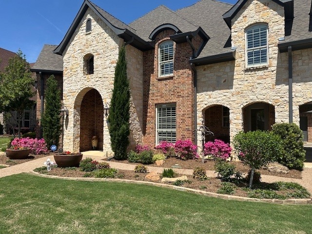 french country home with a shingled roof, stone siding, and a front lawn