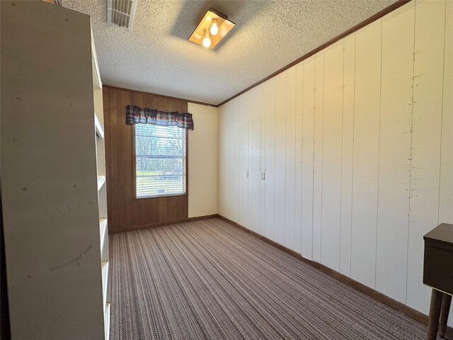 carpeted empty room featuring a textured ceiling, wooden walls, visible vents, baseboards, and ornamental molding