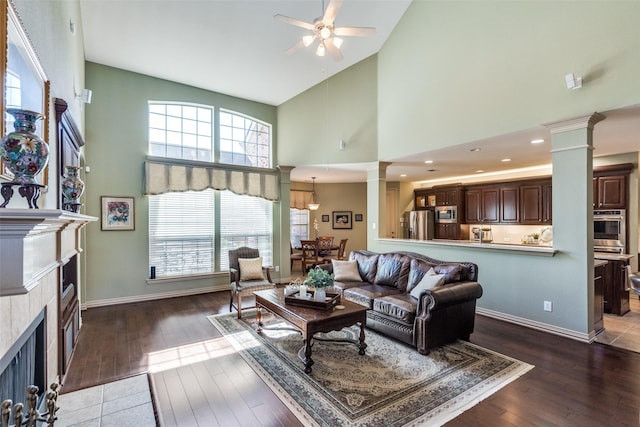 living area with decorative columns, a tiled fireplace, ceiling fan, wood finished floors, and baseboards