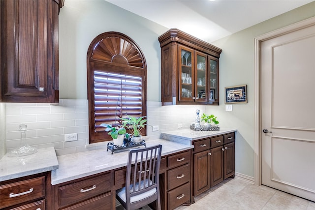 kitchen featuring glass insert cabinets, light tile patterned flooring, built in study area, dark brown cabinets, and light stone countertops