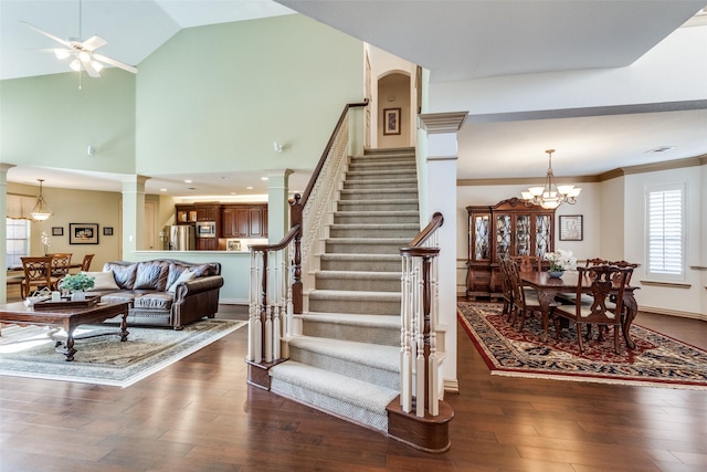 stairs featuring high vaulted ceiling, ceiling fan with notable chandelier, wood finished floors, decorative columns, and crown molding