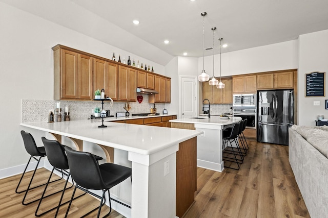 kitchen featuring a center island with sink, appliances with stainless steel finishes, brown cabinetry, under cabinet range hood, and a kitchen bar