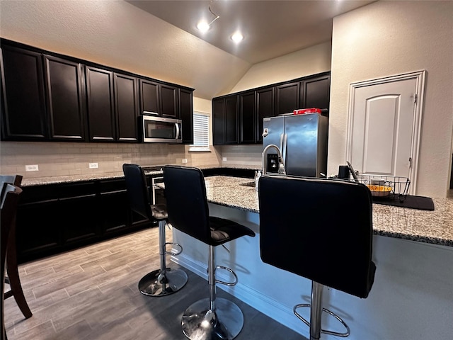 kitchen featuring light stone counters, stainless steel appliances, tasteful backsplash, dark cabinets, and a kitchen breakfast bar