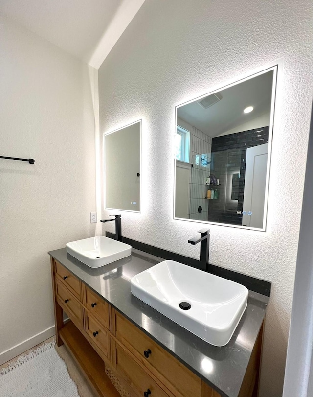 full bath featuring a textured wall, tiled shower, a sink, and double vanity