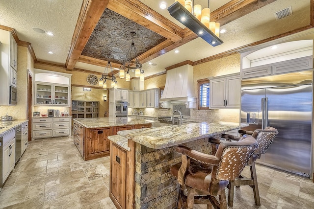 kitchen featuring stainless steel appliances, premium range hood, coffered ceiling, visible vents, and stone tile flooring