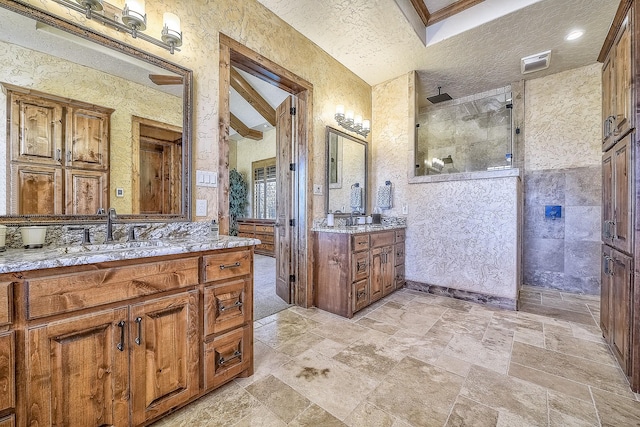 full bathroom featuring a sink, walk in shower, stone tile flooring, a textured ceiling, and two vanities