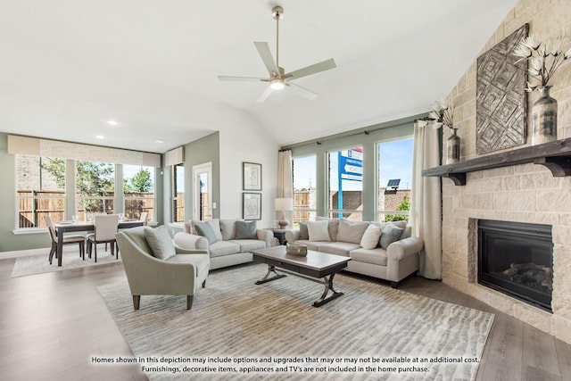 living area featuring a ceiling fan, vaulted ceiling, wood finished floors, and a stone fireplace