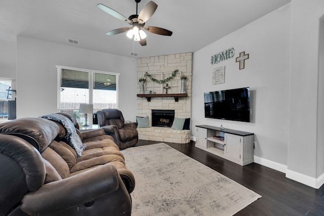 living room featuring dark wood-style flooring, visible vents, a ceiling fan, a stone fireplace, and baseboards