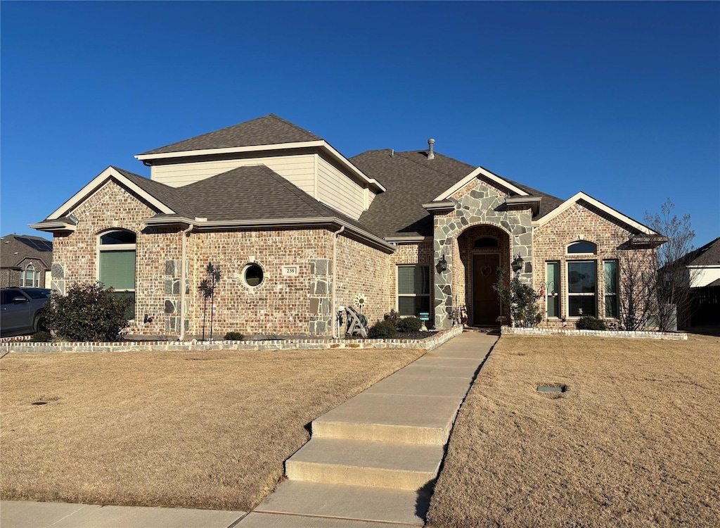 french country style house with stone siding, a shingled roof, and brick siding