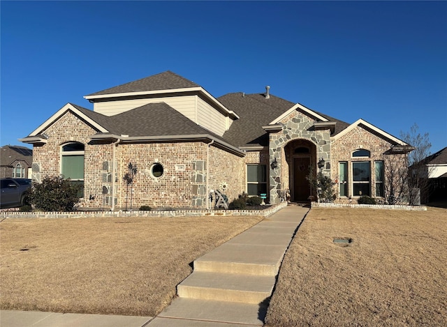 french country style house with stone siding, a shingled roof, and brick siding