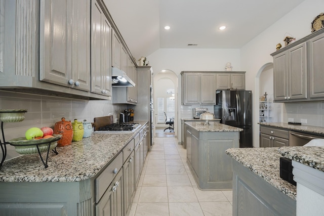 kitchen with arched walkways, appliances with stainless steel finishes, light stone counters, gray cabinets, and under cabinet range hood