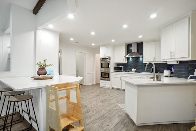 kitchen featuring a peninsula, wall chimney range hood, light wood-style flooring, and stainless steel appliances