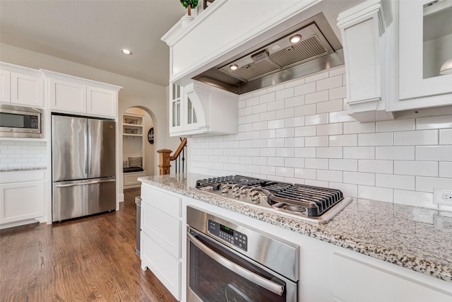 kitchen featuring arched walkways, glass insert cabinets, appliances with stainless steel finishes, premium range hood, and white cabinetry