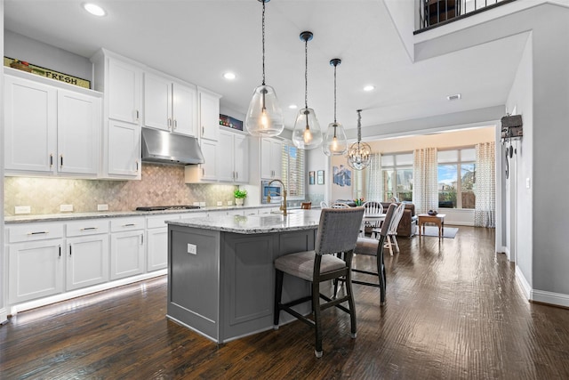 kitchen with a kitchen island with sink, under cabinet range hood, stainless steel gas cooktop, white cabinetry, and decorative backsplash