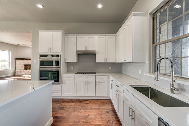 kitchen with white cabinets, dark wood-style floors, stainless steel double oven, under cabinet range hood, and a sink