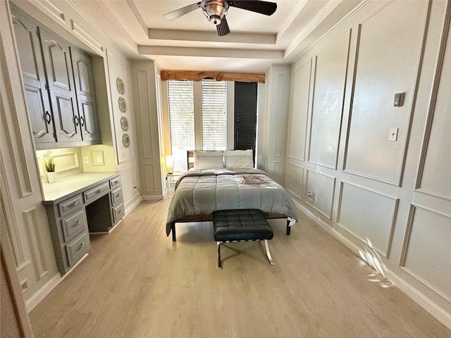 bedroom with a raised ceiling, a decorative wall, light wood-style floors, a ceiling fan, and built in study area