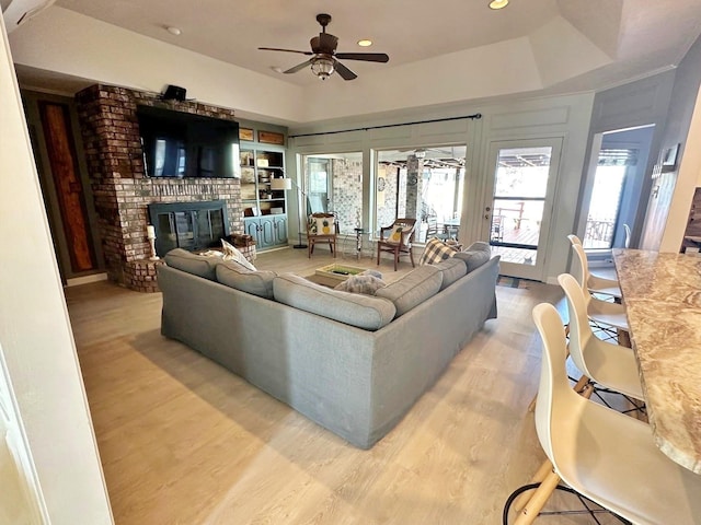 living room featuring a ceiling fan, a tray ceiling, light wood-type flooring, a brick fireplace, and recessed lighting