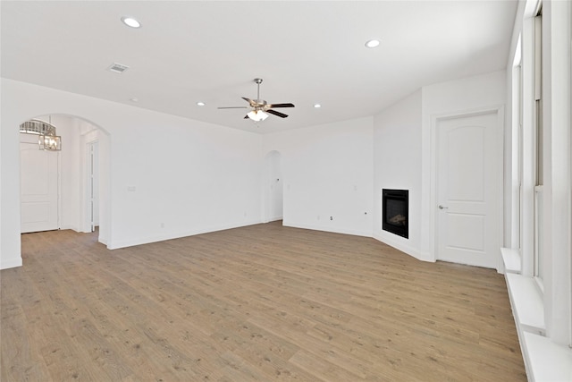 unfurnished living room with arched walkways, ceiling fan with notable chandelier, light wood-type flooring, and a glass covered fireplace