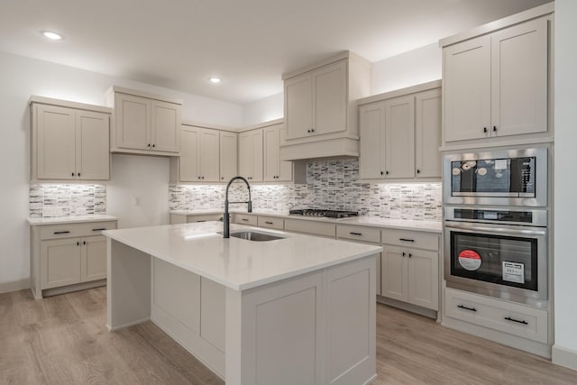 kitchen featuring stainless steel appliances, light countertops, a kitchen island with sink, a sink, and light wood-type flooring