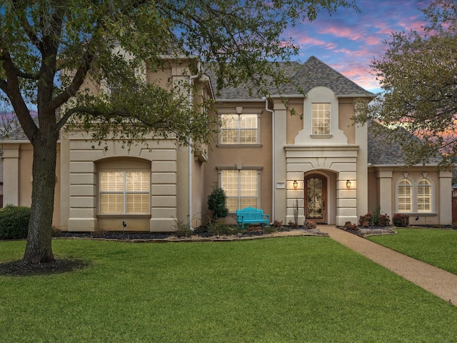 french country style house with a shingled roof, a front lawn, and stucco siding