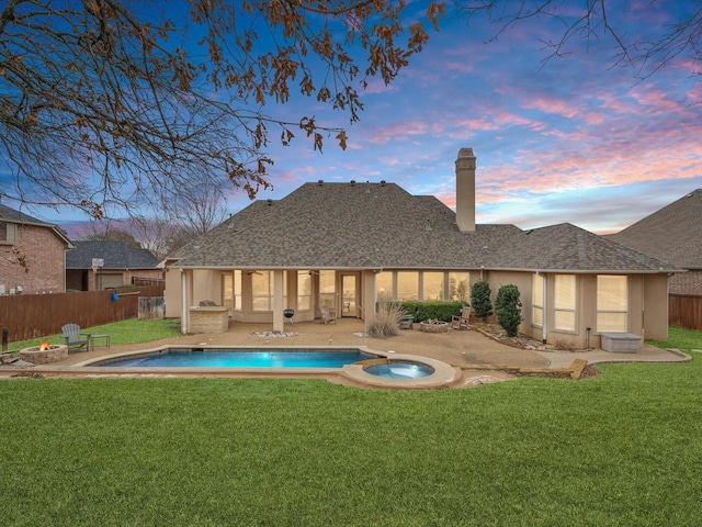 back of house at dusk with fence, a yard, a fire pit, and an in ground hot tub