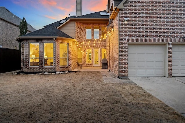 back of property at dusk with a garage, brick siding, concrete driveway, roof with shingles, and a chimney