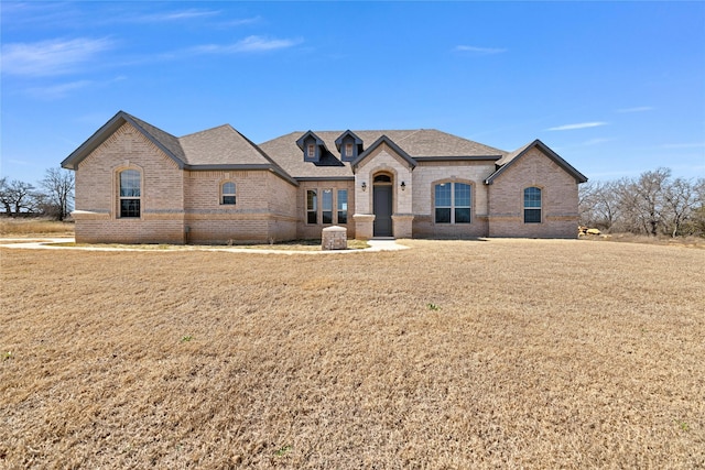 french country inspired facade with a shingled roof, brick siding, and a front lawn