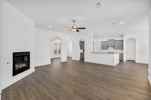 unfurnished living room with dark wood-type flooring, a glass covered fireplace, ceiling fan, and arched walkways