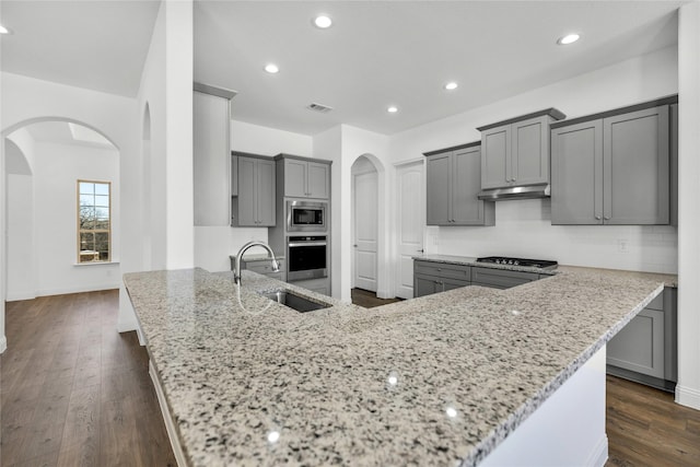 kitchen with arched walkways, gray cabinets, appliances with stainless steel finishes, a sink, and light stone countertops