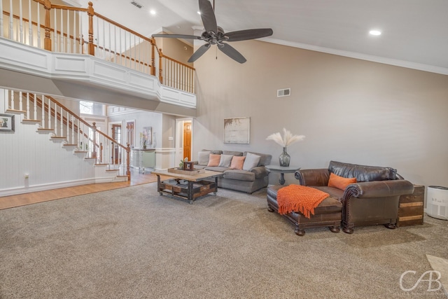 carpeted living room with a towering ceiling, visible vents, stairs, a ceiling fan, and ornamental molding