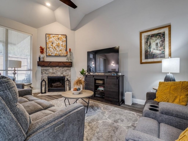 living room with ceiling fan, a stone fireplace, wood finished floors, baseboards, and vaulted ceiling