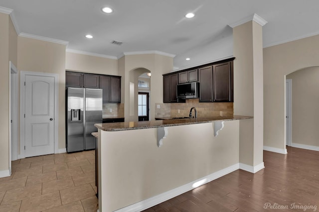 kitchen featuring dark brown cabinetry, arched walkways, dark stone countertops, a peninsula, and stainless steel appliances