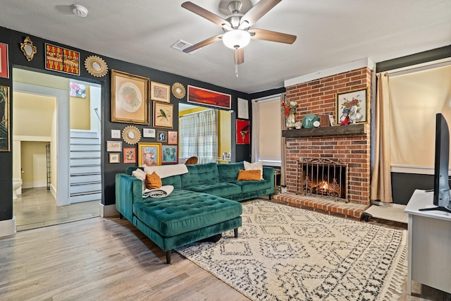 living area featuring ceiling fan, wood finished floors, visible vents, stairway, and a brick fireplace