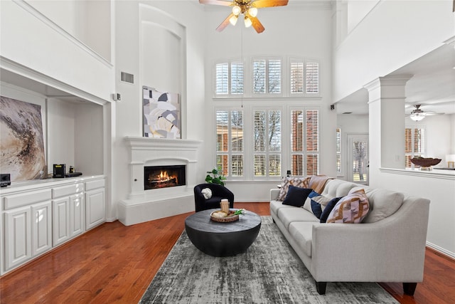 living room with ceiling fan, wood finished floors, visible vents, a lit fireplace, and decorative columns