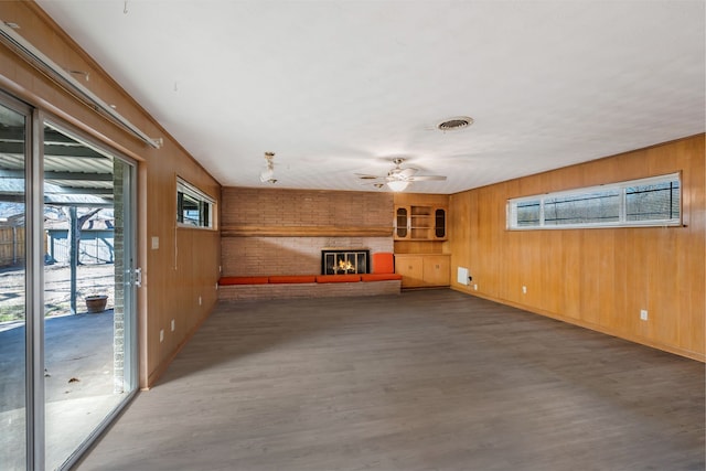 unfurnished living room featuring ceiling fan, wood walls, wood finished floors, visible vents, and a brick fireplace