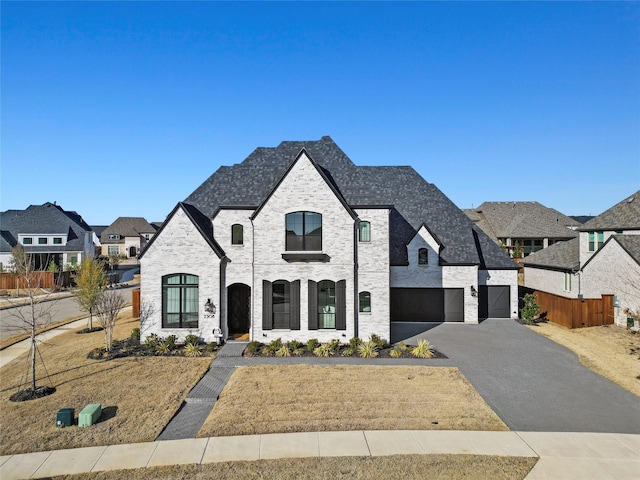 french country inspired facade featuring brick siding, an attached garage, fence, a residential view, and driveway