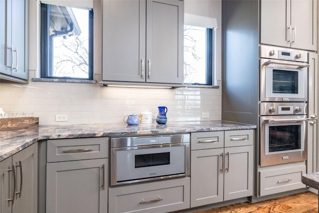 kitchen featuring light wood finished floors, decorative backsplash, light stone countertops, gray cabinets, and stainless steel double oven