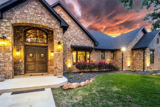 exterior entry at dusk with a shingled roof, stone siding, and a yard