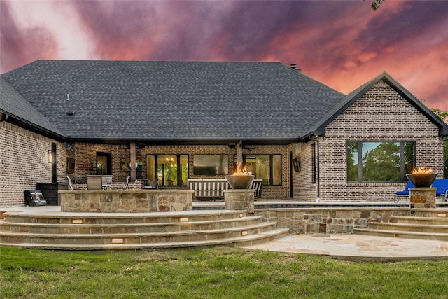 back of property at dusk with a patio, brick siding, roof with shingles, and a lawn