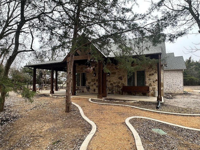 rear view of house with stone siding, a patio area, and roof with shingles