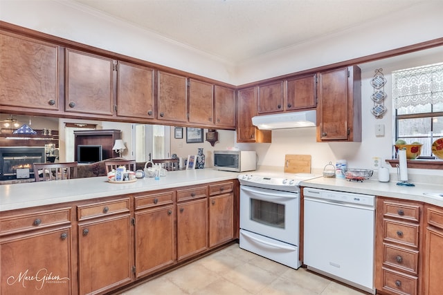 kitchen with light countertops, brown cabinetry, a warm lit fireplace, white appliances, and under cabinet range hood