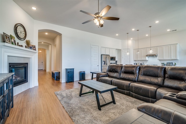 living room featuring light wood finished floors, arched walkways, a tiled fireplace, ceiling fan, and recessed lighting